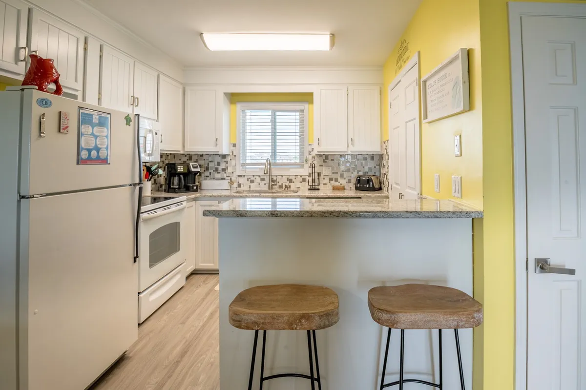 A view of a modern kitchen with granite countertops, sleek stools for bar seating, vibrant yellow walls, and a window letting in natural light. The space feels fresh, functional, and tailor-made for cooking and socializing.