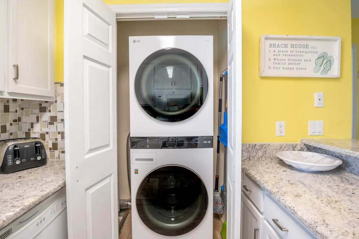 A functional laundry nook with a stacked washer and dryer, granite countertop, toaster, and a lighthearted decorative sign. The area is conveniently integrated into the living space, making extended stays and family trips easier.