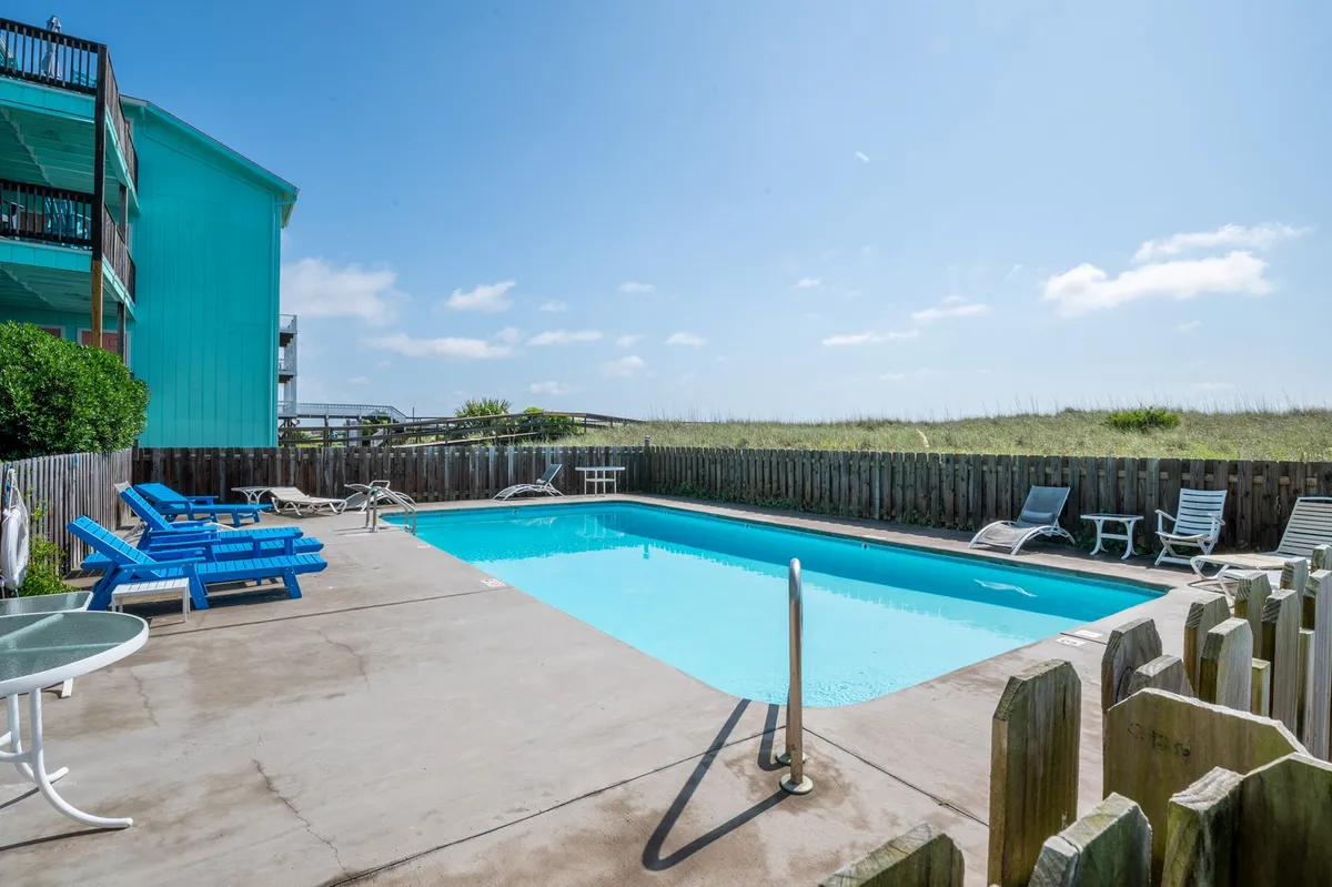 This image captures a vibrant outdoor swimming pool surrounded by green foliage and bordered by a fence. Lounge chairs are arranged poolside, suggesting a setting perfect for relaxation or family enjoyment. The atmosphere is leisurely, emphasizing poolside activities such as sunbathing, swimming, and relaxing outdoors.