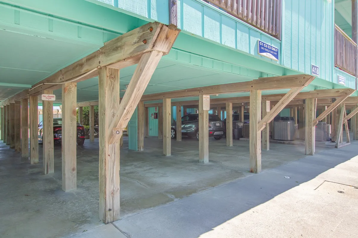 An open parking area beneath a building with colorful facades and sturdy wooden pillars. The scene shows ample space for vehicles and an accessible layout, reflecting a casual and relaxed mood.