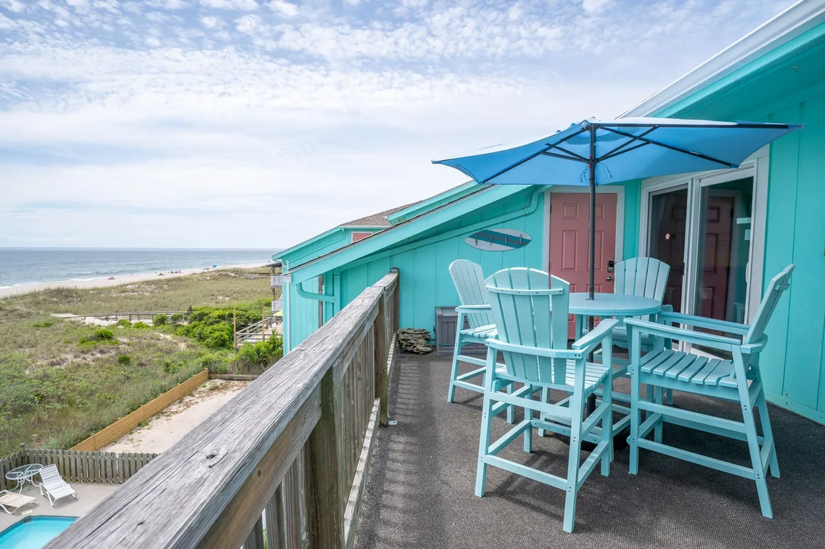 An inviting outdoor scene highlighting the turquoise building, an outdoor seating area with tables and chairs, a view of the beach, and a sparkling pool. The space is perfect for social gatherings, outdoor meals, or relaxing with friends while enjoying ocean views.
