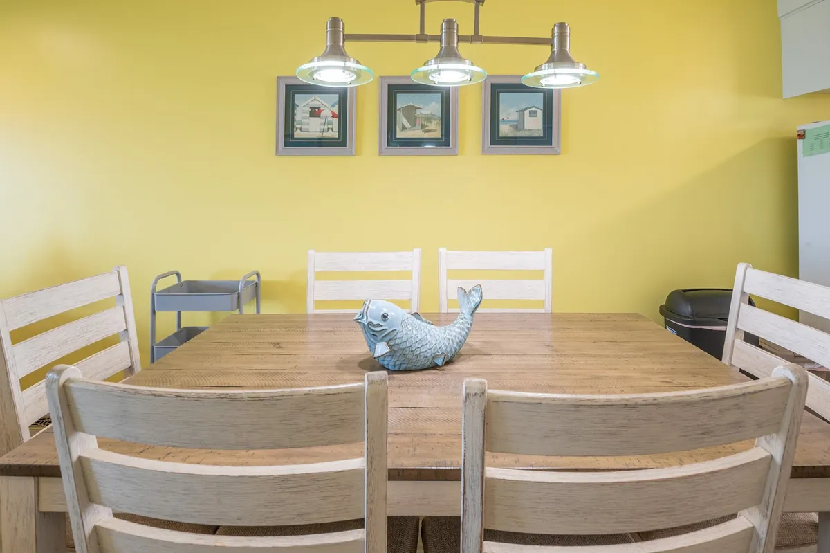 A wood dining table set with a playful blue fish centerpiece, surrounded by comfortable seating and framed by colorful artwork. The space is inviting, ideal for shared meals and lively conversation.