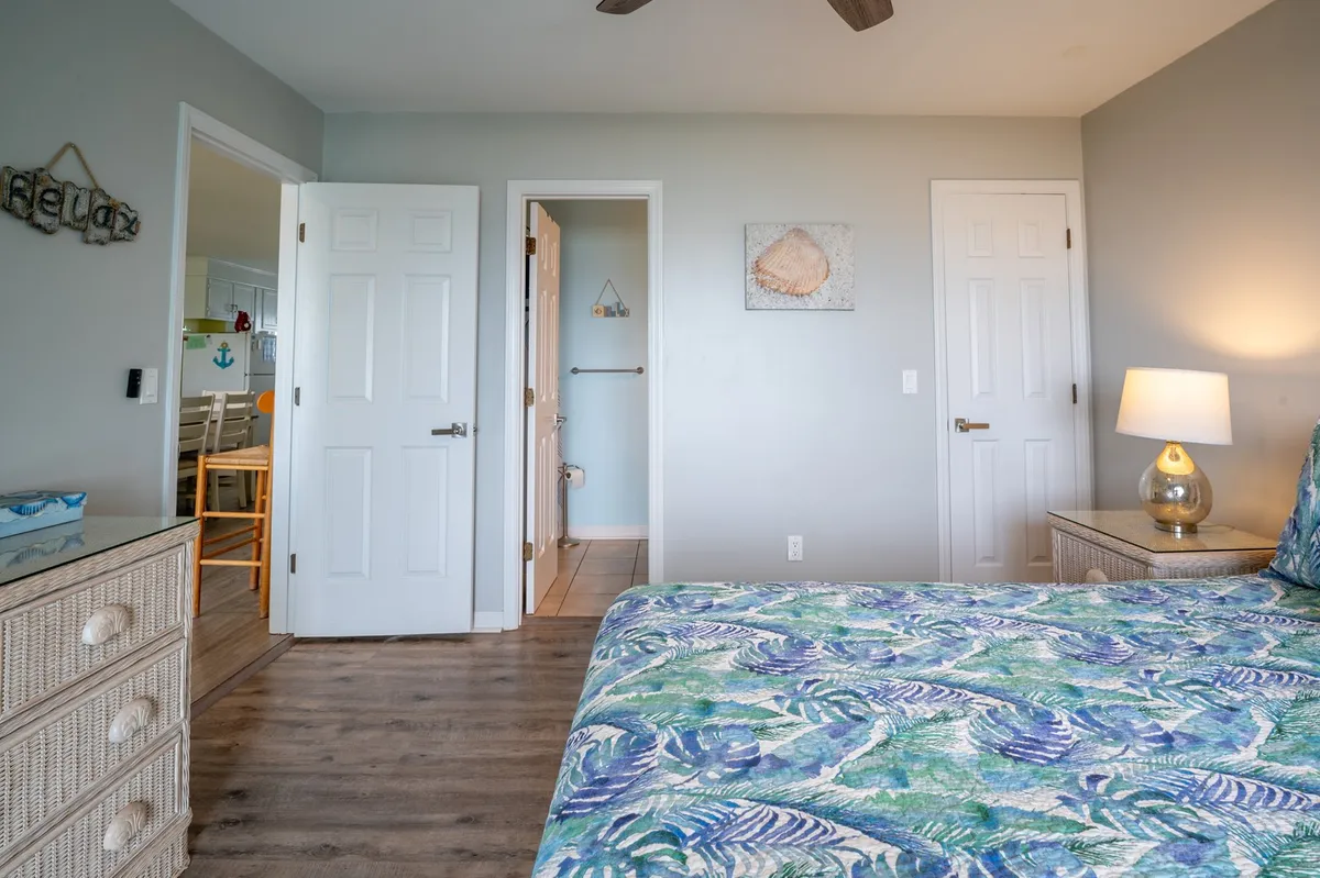 A tranquil bedroom scene with a bed dressed in tropical-patterned bedding, a nightstand with a lamp, and open doors leading to adjacent rooms. Sunlight streams in, creating a calm, restful environment ideal for unwinding after a day at the beach.