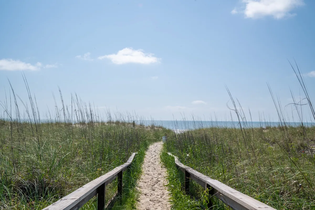 This exterior image features a wooden walkway leading through sand dunes and grass toward the ocean, beneath a wide, blue sky. The path invites viewers to imagine a sunrise stroll or sunset walk, connecting the property directly to the beach.