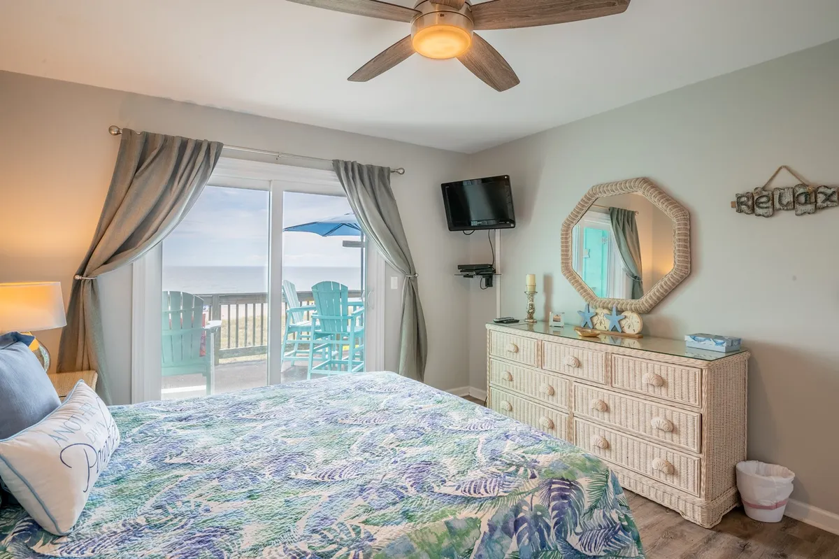 A tranquil primary bedroom with a plush bed dressed in patterned bedding, a dressing table with a decorative mirror, and sliding glass doors framing a beautiful ocean view. Soft natural light fills the space, and the doors open directly to the private deck, inviting sea breezes inside.