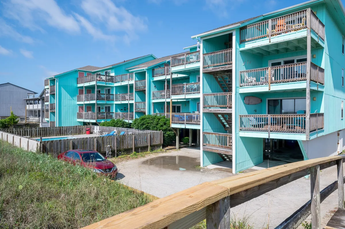 A wider exterior view of the turquoise facade, with multiple balconies, lush landscaping, and a glimpse of the parking area. The scene suggests a quiet, friendly environment and highlights opportunities for outdoor relaxation and socializing.