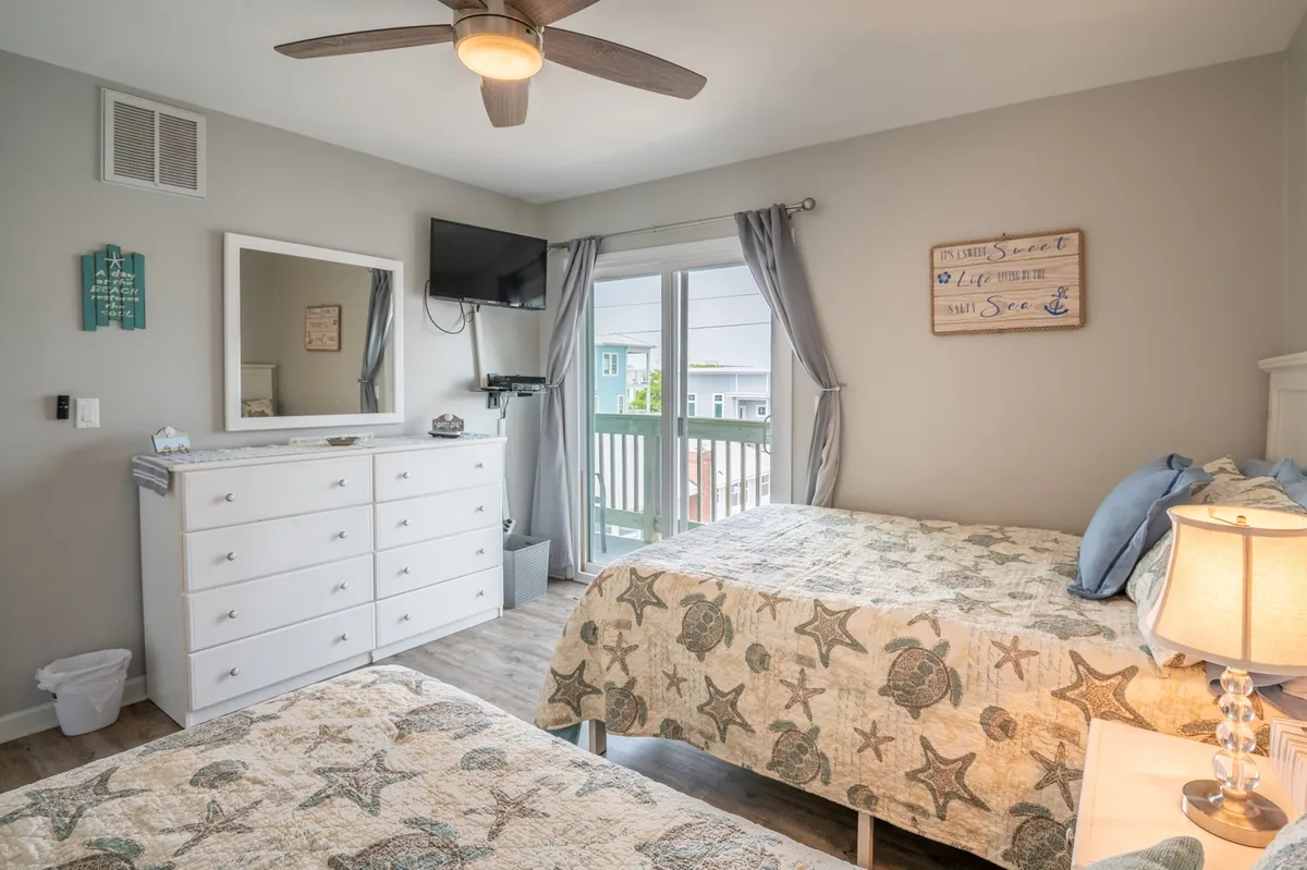 The guest bedroom features two beds with starfish-patterned quilts, a mirrored dresser, and large balcony doors that allow natural light to flood the room. The private balcony beyond offers a spot to enjoy morning coffee or a quiet evening drink while taking in the island views.