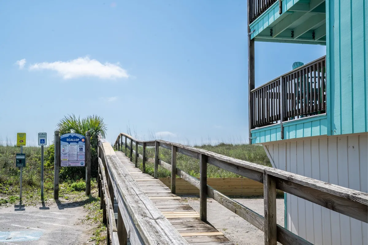 This image features a wooden walkway leading directly from the property towards the sandy beach, with a beach access sign, palm tree, and a glimpse of the building's balcony. The walkway invites exploration and highlights the convenience of private, direct beach access.