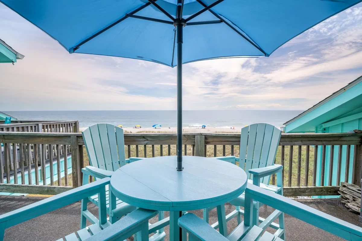 A tranquil oceanfront deck set with blue Adirondack chairs and a blue umbrella, facing an expansive ocean view. The setting is ideal for enjoying sunrise coffee, afternoon relaxation, or evening cocktails while listening to the waves.