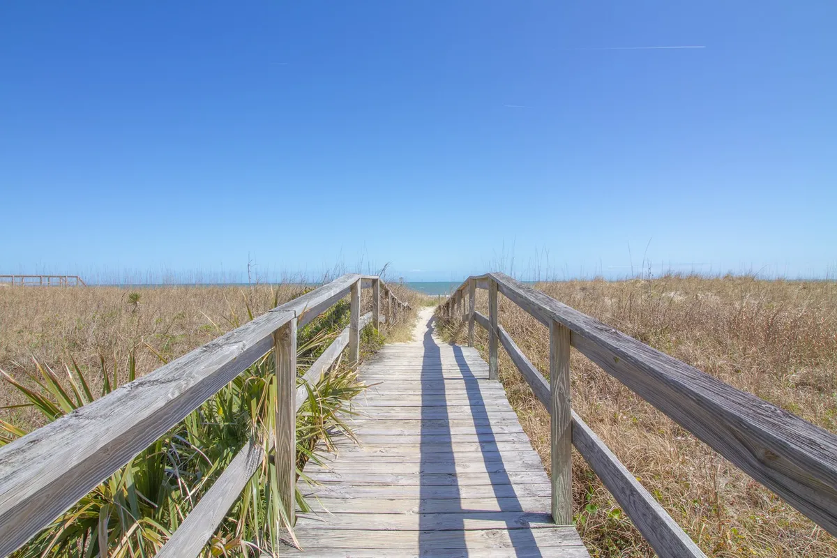A long, wooden boardwalk stretches through beach grass toward a sweeping ocean view. The path invites guests to walk directly onto the sandy shore, with tranquil water and open skies in the background. The mood is peaceful, evoking early morning or sunset strolls.
