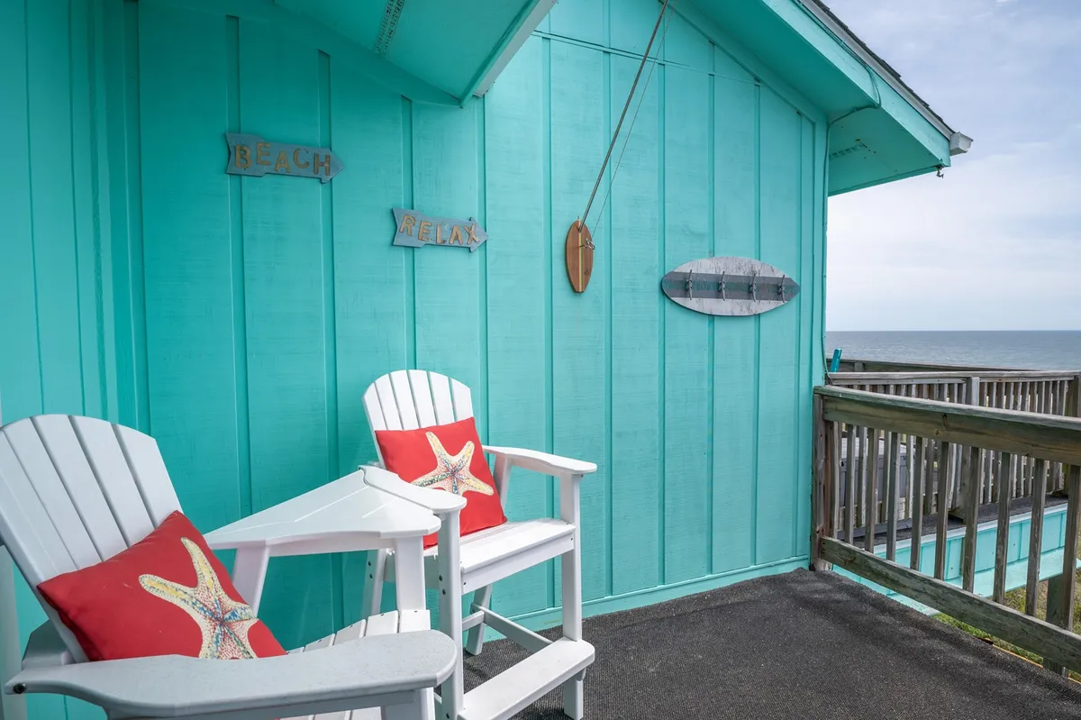 An inviting exterior seating area with white chairs and a starfish pillow facing the ocean, turquoise walls, and beach-themed signs saying 'BEACH' and 'RELAX.' The setup is ideal for enjoying a sunrise coffee, reading a book, or taking in the sound of the waves.