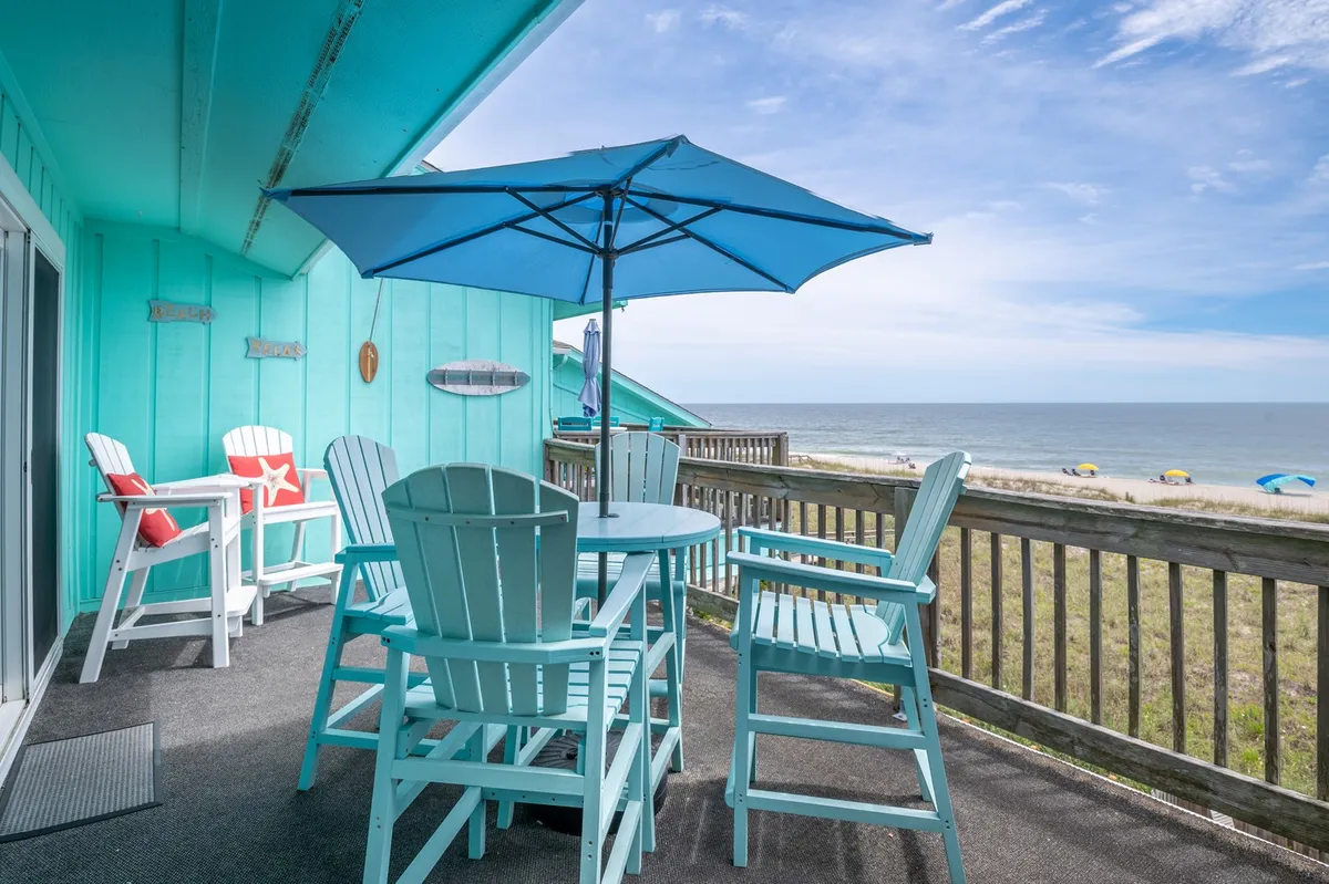 The image showcases a private balcony set with a table and chairs beneath a vibrant blue umbrella, overlooking a pristine stretch of sandy Carolina Beach and endless ocean views. The inviting scene suggests a perfect spot for enjoying meals, relaxing with coffee at sunrise, or sharing sunset drinks with friends as waves roll in below.
