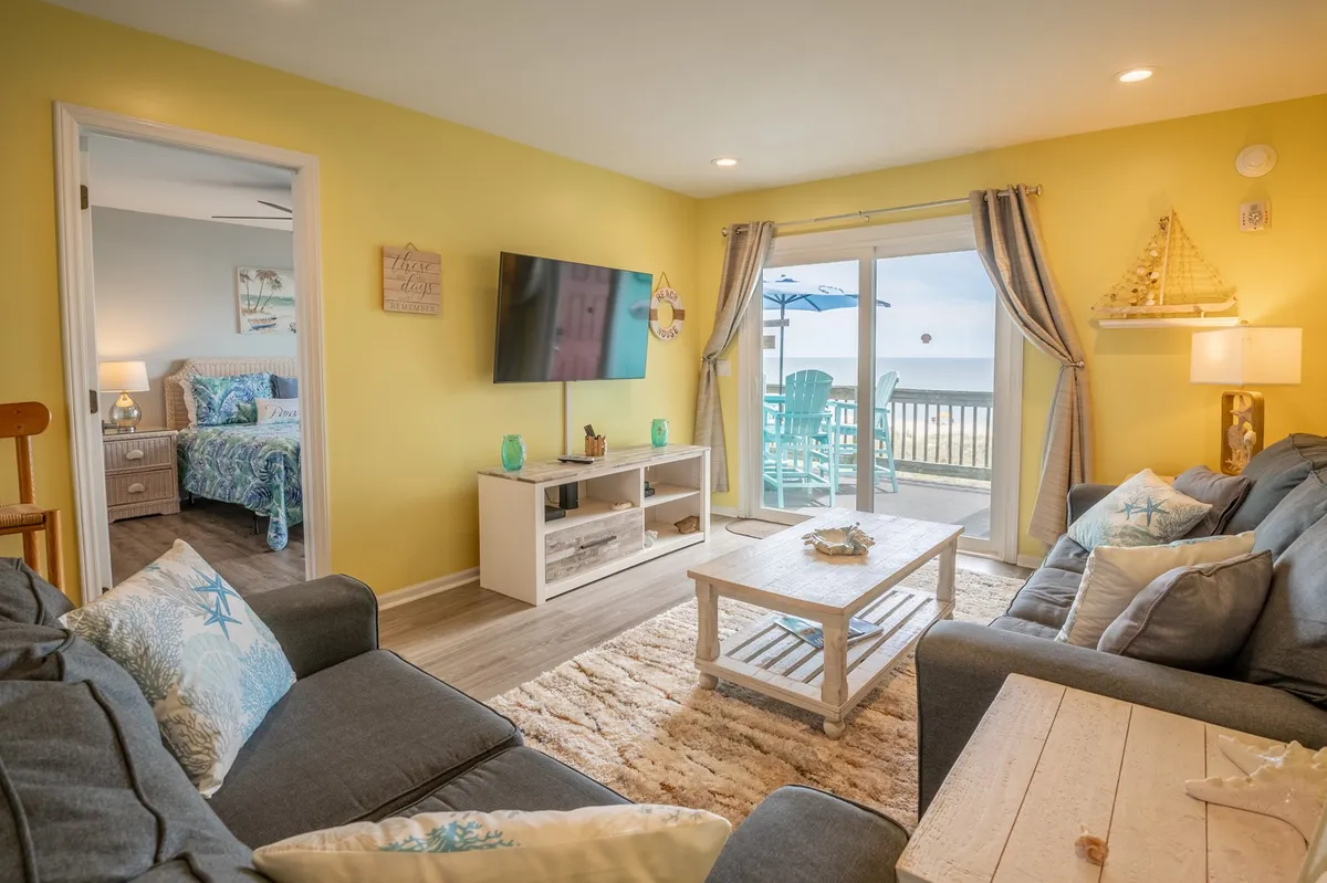A comfortable living space featuring a sofa with decorative pillows, a coffee table adorned with shell decor, a television, and large sliding glass doors that open onto an oceanfront deck. Coastal sunlight pours in, illuminating the room and offering seamless access to outdoor views.